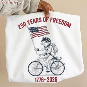 Könnte beinhalten: Weißes Sweatshirt mit einer Grafik einer Froschfigur in Kolonialkleidung, die Fahrrad fährt und eine amerikanische Flagge hält. Der Text "250 YEARS OF FREEDOM" und "1776-2026" sind rot gedruckt.