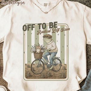 Puede incluir: Camiseta blanca con una ilustración caprichosa. Una rana con gorra y suéter monta en bicicleta con un gato en una cesta. El texto dice "OFF TO BE Radical Left Scum."