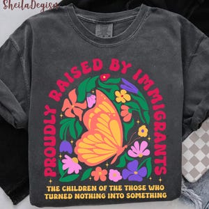 May include: Dark gray sweatshirt with a colorful graphic. The design features a butterfly, flowers, and text that reads "PROUDLY RAISED BY IMMIGRANTS" and "THE CHILDREN OF THE THOSE WHO TURNED NOTHING INTO SOMETHING."