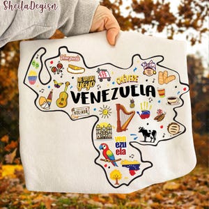 May include: A white, rectangular textile featuring a map of Venezuela with colorful illustrations and text. The word "VENEZUELA" is prominently displayed. Various icons include a guitar, harp, and a parrot. The text "ME VADAR yeyo" and "CHEVERE" are also visible.
