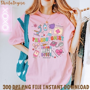 Puede incluir: Camiseta rosa claro con un gráfico colorido de "Flebotomista" que presenta herramientas médicas y el texto "I stab people". El diseño incluye un microscopio, una mascarilla médica y un corazón con una línea de latido.