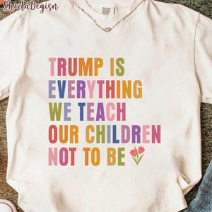 May include: Cream-colored t-shirt with the text "TRUMP IS EVERYTHING WE TEACH OUR CHILDREN NOT TO BE" in colorful block letters. Two small pink flowers with green stems are at the bottom right of the text.