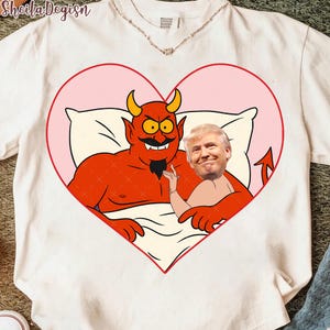 Op de afbeelding: Wit T-shirt met een cartoonduivel in bed met het gezicht van een persoon in een roze hart. De duivel heeft hoorns en een lachend gezicht. Het shirt heeft bovenaan de tekst "Sheila Degisn".