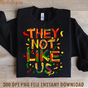Op de afbeelding: Zwarte sweatshirt met de tekst "THEY NOT LIKE US" in kleurrijke, gestileerde letters. Het ontwerp bevat rode, gele, groene en blauwe accenten. De woorden zijn in een opvallend lettertype.