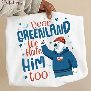 Pode incluir: Camisola branca com o texto "Dear Greenland We Hate Him Too" em azul e vermelho. Um urso polar de desenho animado usando um moletom azul e um gorro vermelho é apresentado no lado direito da camisola.