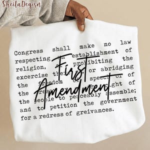 Puede incluir: Tela blanca con el texto de la Primera Enmienda en negro. Las palabras "First Amendment" están en una fuente grande y estilizada. El texto incluye frases como "El Congreso no promulgará ninguna ley" y "libertad de expresión".