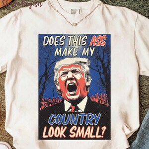 May include: White t-shirt featuring a graphic with the text "DOES THIS ASS MAKE MY COUNTRY LOOK SMALL?" The graphic depicts a man with red and white facial features, set against a blue background with silhouettes of trees and a crowd.