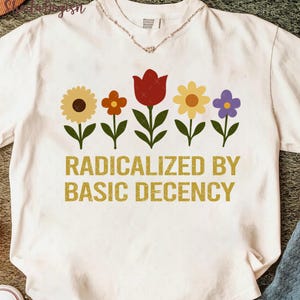 Op de afbeelding: Wit T-shirt met een bloemenontwerp. Het ontwerp toont vijf kleurrijke bloemen en de tekst "RADICALIZED BY BASIC DECENCY" in goud. De bloemen zijn onder meer een zonnebloem, een oranje bloem, een rode tulp, een gele bloem en een paarse bloem.