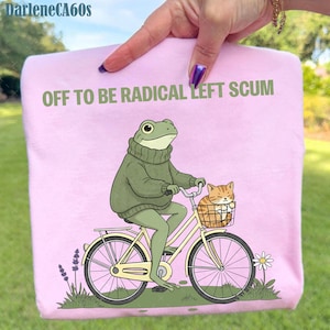 Könnte beinhalten: Rosa Sweatshirt mit dem Text "OFF TO BE RADICAL LEFT SCUM". Die Grafik zeigt einen Frosch, der Fahrrad fährt, mit einer Katze im Korb. Der Frosch trägt einen grünen Pullover. Das Fahrrad ist cremefarben.