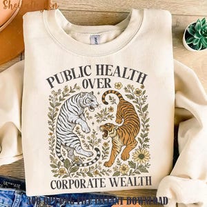 Op de afbeelding: Een crèmekleurig sweatshirt met een grafisch ontwerp met twee tijgers in een bloemig kader. De tekst "Openbare gezondheid boven bedrijfsvermogen" staat op het sweatshirt gedrukt.
