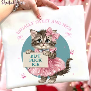 Puede incluir: Cojín cuadrado blanco con una ilustración de un gatito de dibujos animados. El gatito lleva un vestido y un lazo rosas, sosteniendo un cartel que dice "BUT FUCK ICE". El texto "USUALLY SWEET AND NICE" está arqueado sobre el gatito.
