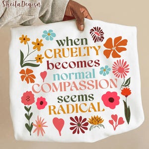May include: White sweatshirt featuring the quote "When cruelty becomes normal, compassion seems radical" in multiple colours. The design incorporates floral illustrations and a heart shape.