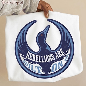 Pode incluir: Camisola branca com um emblema de pássaro azul marinho e azul claro dentro de um círculo. O texto "REBELLIONS ARE BUILT ON HOPE" é branco. A marca "SheilaDegisn" é visível no canto superior esquerdo.