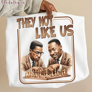 May include: White sweatshirt featuring a brown framed graphic with the text "THEY NOT LIKE US". The graphic depicts two men playing chess. The sweatshirt is held by a person.
