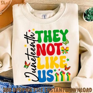 Peut inclure: Sweat-shirt crème avec l'inscription "Juneteenth" en écriture noire et "THEY NOT LIKE US" en lettres colorées. Le motif comprend des papillons, des étoiles et des poings levés rouges, jaunes, verts et noirs.
