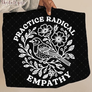 May include: Black square textile with a white floral and bird design. The text "PRACTICE RADICAL EMPATHY" is arranged around the design. The design features a bird perched on a branch with flowers and leaves.