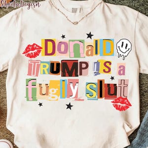Op de afbeelding: Wit t-shirt met een kleurrijk grafisch ontwerp. Het ontwerp bevat de woorden "Donald Trump is a fugly slut" in een losgeldschriftstijl, samen met rode lippen, sterren en een smeltend smiley-gezicht.