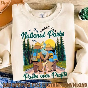 Peut inclure: Un sweat-shirt beige avec un motif graphique de deux ours portant des chapeaux de rangers et des sacs &agrave; dos. Les ours marchent sur un sentier dans une for&ecirc;t avec le texte "Prot&eacute;gez nos parcs nationaux" et "Les parcs avant les profits".