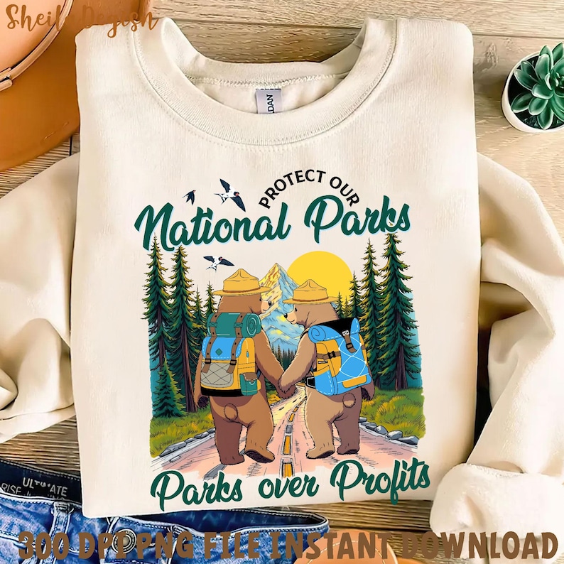 Peut inclure: Un sweat-shirt beige avec un motif graphique de deux ours portant des chapeaux de rangers et des sacs &agrave; dos. Les ours marchent sur un sentier dans une for&ecirc;t avec le texte "Prot&eacute;gez nos parcs nationaux" et "Les parcs avant les profits".
