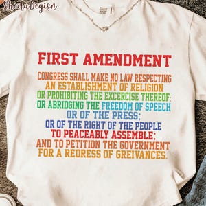 Puede incluir: Camiseta blanca con el texto "FIRST AMENDMENT" en rojo, seguido del texto de la enmienda en colores del arcoíris. La camiseta tiene cuello en V y está hecha de un material suave.