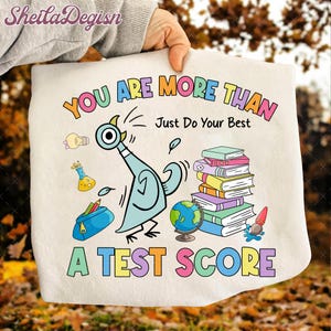 May include: A white tote bag with a colorful cartoon design. The design features a blue pigeon, books, a globe, and the text "YOU ARE MORE THAN A TEST SCORE. Just Do Your Best."