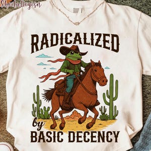 以下が含まれることがあります： 白いTシャツ。カウボーイハットをかぶったカエルの漫画が茶色の馬に乗っています。カエルは緑色のジャケットとスカーフを着ています。「RADICALIZED by BASIC DECENCY」の文字。背景にはサボテンと砂漠の風景があります。