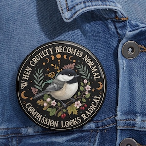 May include: A round pinback button featuring a black-capped chickadee perched on a branch with leaves and flowers. The black background includes moon phases and the text: WHEN CRUELTY BECOMES NORMAL, COMPASSION LOOKS RADICAL. The button is attached to blue denim.