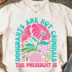 Puede incluir: Camiseta blanca con un diseño floral y el texto "IMMIGRANTS ARE NOT CRIMINALS THE PRESIDENT IS" en turquesa y rosa. El diseño presenta flores rosas, hojas turquesas y una regadera rosa.