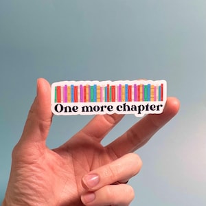 May include: A rectangular sticker featuring a colorful row of books. The sticker has the text "One more chapter" in a black font. The books are in various colors, including red, green, yellow, and purple. The sticker is held by a hand against a light blue background.