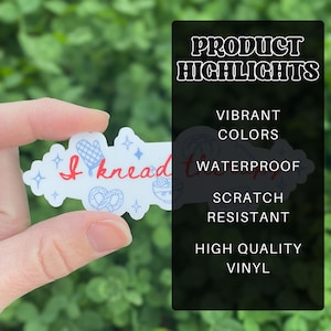May include: A white sticker with the text "I knead therapy" in red script, with blue illustrations of a mitten, pretzel, and other baking-related items. The sticker is held in a hand against a green background.