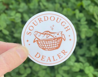 May include: A circular white sticker with an orange border and orange text that reads SOURDOUGH DEALER. The center features an orange line drawing of a basket filled with loaves of bread, accented by small four-pointed star symbols.