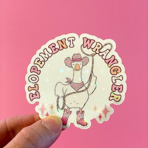 May include: A sticker featuring a cartoon goose dressed as a cowgirl, wearing a pink hat and boots, holding a lasso. The sticker has the text "Elopement Wrangler" in a pink, circular design with star accents.