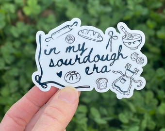 May include: A white die-cut sticker featuring dark blue line art illustrations of baking tools including a rolling pin, whisk, apron, and various breads. The text in the center reads in my sourdough era, accompanied by a small heart icon.