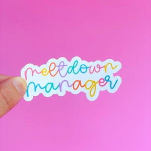May include: A white sticker with the words "meltdown manager" in colorful, handwritten-style lettering. The sticker is held against a bright pink background. The text is outlined in various colors, including orange, blue, and yellow.