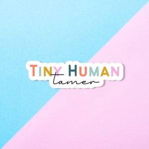 May include: A white sticker with the text "TINY HUMAN tamer" in colorful letters on a pink and blue background.