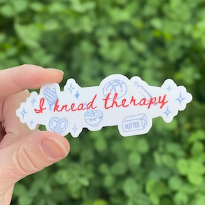 May include: A white sticker with the text "I knead therapy" in red script. The sticker features blue illustrations of baking tools and food items, including a rolling pin, bread, and a bowl. The sticker is held against a green background.