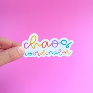 May include: A colorful sticker with the words "chaos coordinator" in a rainbow script font. The sticker has a white border and is held against a bright pink background.