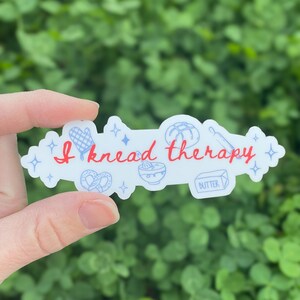 May include: A white sticker with the text "I knead therapy" in red script. The sticker features illustrations of baking tools and food items, including a pretzel, rolling pin, and butter.