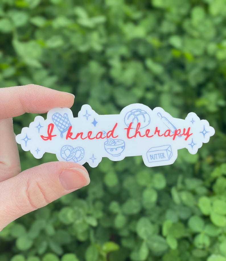 May include: A white sticker with the text "I knead therapy" in red script. The sticker features illustrations of baking tools and food items, including a pretzel, rolling pin, and butter.