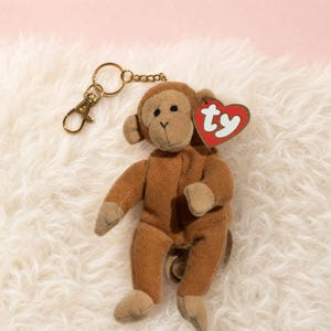May include: A brown plush monkey keychain featuring a soft fabric body, small ears, and a light brown face. A gold-toned metal lobster clasp and chain are attached to the top. A red heart-shaped tag with the white text ty is attached to the ear.