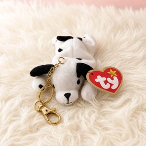May include: A plush dalmatian dog keychain featuring a white body with black spots, black ears, and a black nose. A gold-toned metal lobster clasp and ring are attached to the top. A heart-shaped red tag with the white text ty is attached.