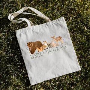 May include: A white canvas tote bag with a brown bear, raccoon, fox, deer, and rabbit printed on it. The text "Respect the Locals" is printed below the animals.