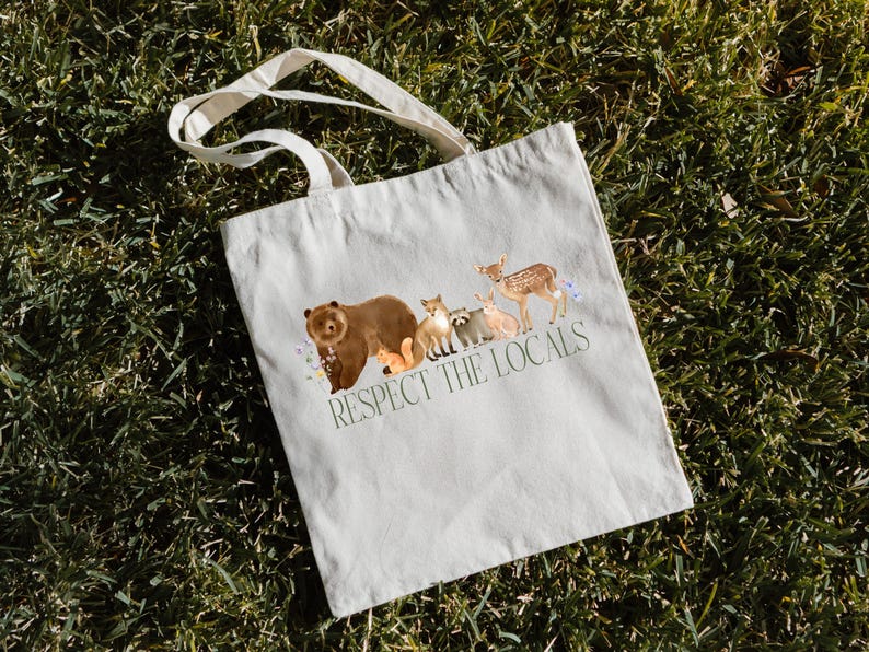 May include: A white canvas tote bag with a brown bear, raccoon, fox, deer, and rabbit printed on it. The text "Respect the Locals" is printed below the animals.