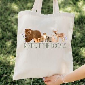May include: A white canvas tote bag with a brown bear, fox, raccoon, deer, and squirrel printed on it. The bag has the text "Respect the Locals" printed on it.