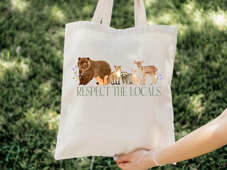 May include: A white canvas tote bag with a brown bear, fox, raccoon, deer, and squirrel printed on it. The bag has the text "Respect the Locals" printed on it.