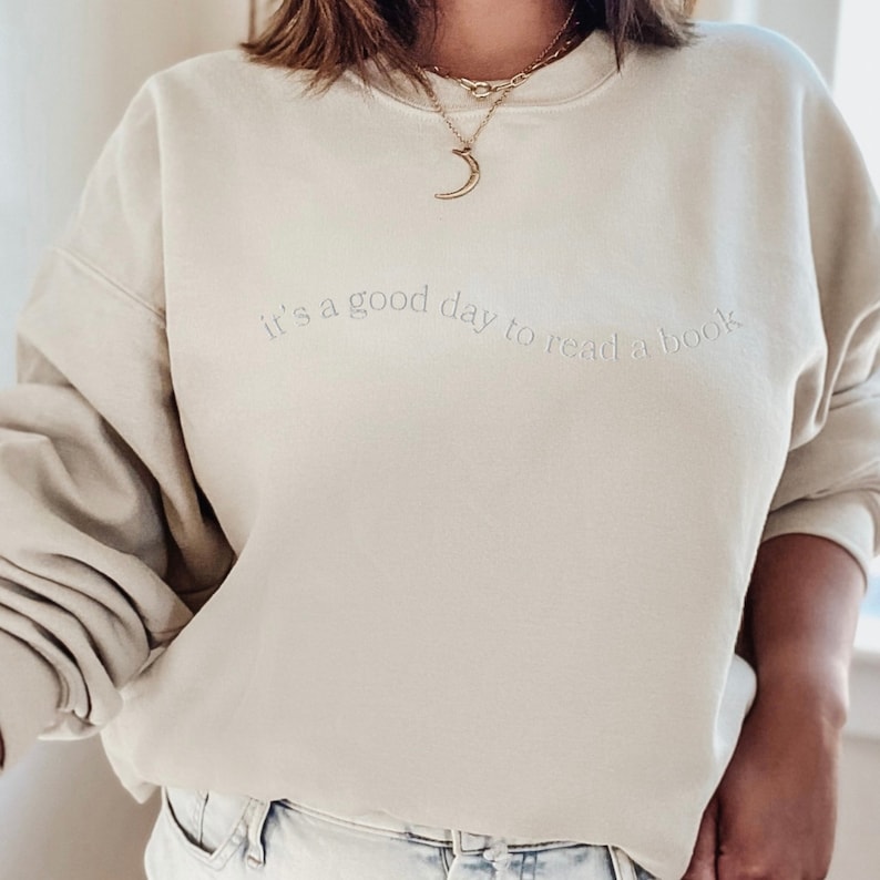 May include: A light beige sweatshirt with the text "it's a good day to read a book" in a light gray color. The sweatshirt has a crew neck and long sleeves.