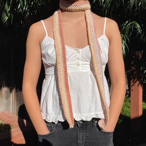 May include: A long, crocheted scarf with stripes of pink, cream, and beige. The scarf is worn around the neck of a person wearing a white top and jeans.
