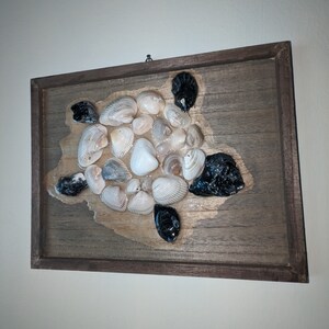 May include: A framed wooden wall art piece featuring a sea turtle made from seashells. The turtle's shell is made from a variety of seashells in different shapes and sizes. The turtle's head and legs are made from black stones.