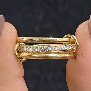 Könnte beinhalten: Ein Goldring bestehend aus drei miteinander verbundenen Ringen. Der mittlere Ring ist mit einer Reihe kleiner, klarer, rund geschliffener Steine besetzt, während die beiden äußeren Ringe aus glattem, poliertem Gold bestehen.