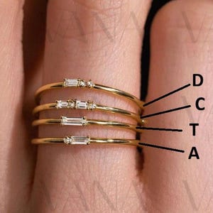 May include: A set of four delicate gold rings, each with a thin band. Three rings feature small, rectangular gemstones, while the fourth has tiny round stones. The rings are stacked on a finger, showcasing their elegant design.